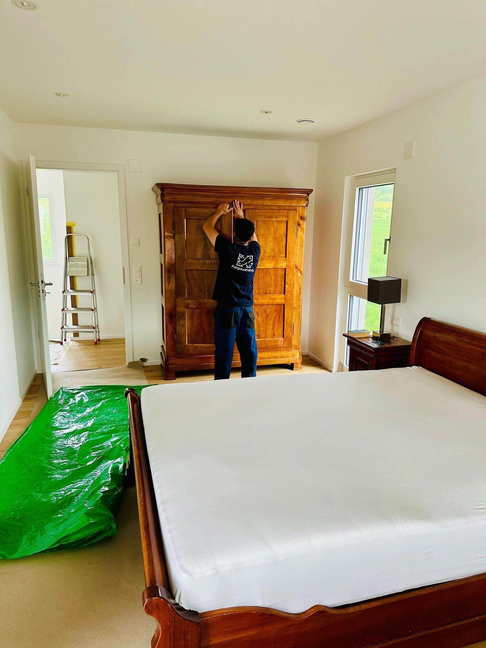 Eine Person steht in einem hellen Schlafzimmer neben einem Holzbett und einem Kleiderschrank; 