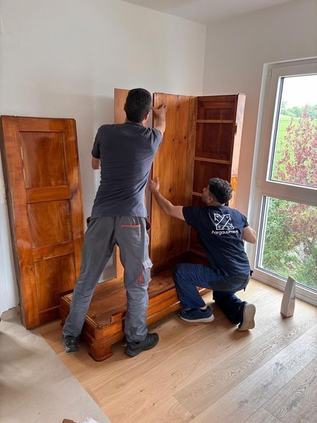 Zwei Personen bauen in einem hellen Zimmer neben einem Fenster einen Holzschrank zusammen.