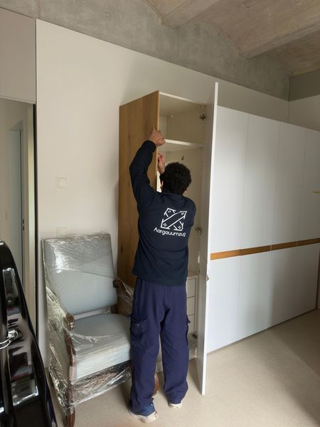 Eine Person steht in einem Raum und montiert einen Holzschrank neben gestapelten Stühlen und einer Schiebetür.