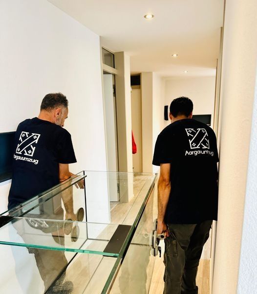 Zwei Arbeiter in schwarzen Hemden gehen einen hellen Flur entlang, der an einem Glasgeländer vorbeiführt.