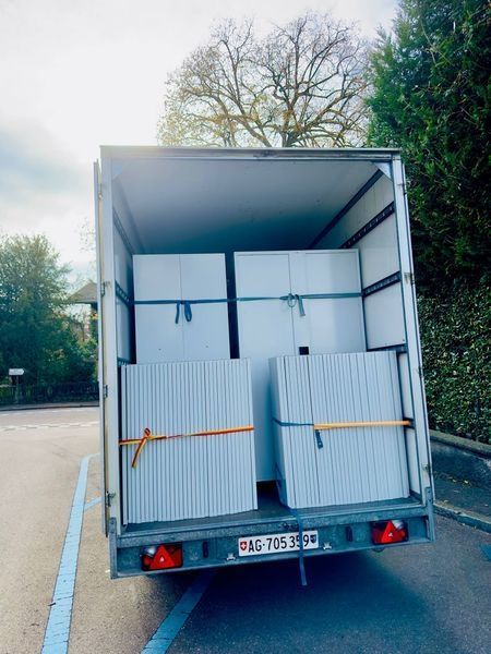 Ein offener Umzugswagen mit weissen Ladeflächen und orangefarbenen Gurten parkt auf einer Strasse.