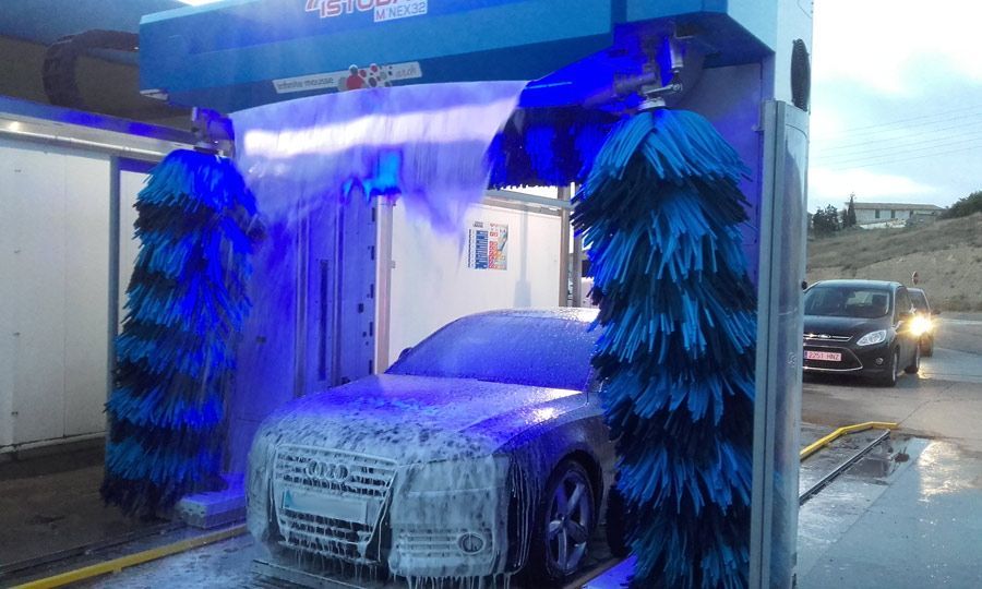 Un coche se lava en un lavadero con cepillos azules y espuma. Un coche espera en la fila afuera.