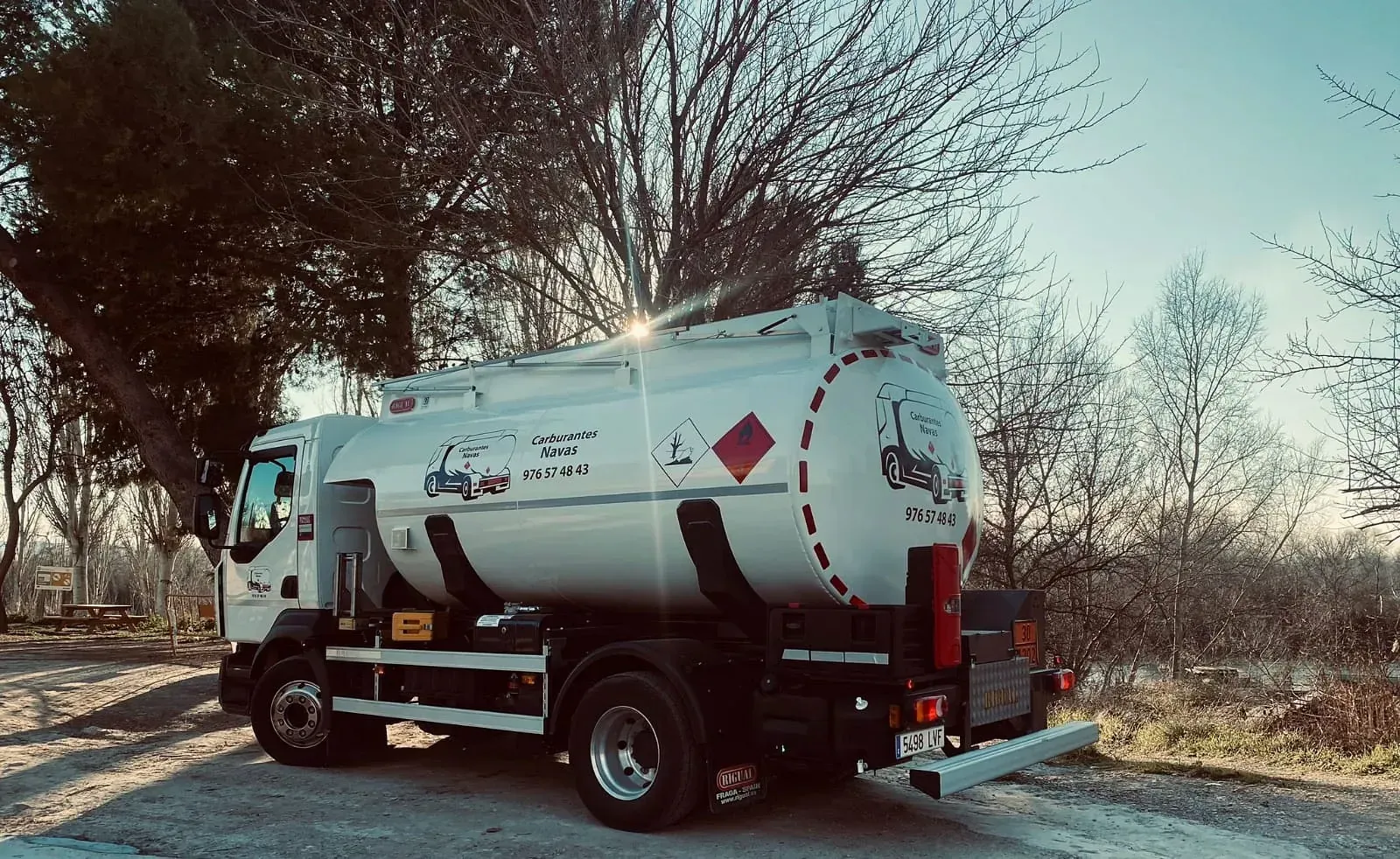 Camión cisterna de combustible de color blanco estacionado al aire libre, cerca de árboles.
