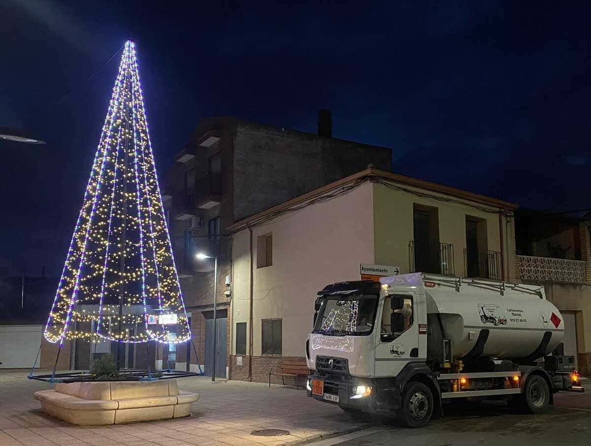 Un árbol de Navidad iluminado junto a un camión blanco con tanque en una calle adoquinada por la noche.