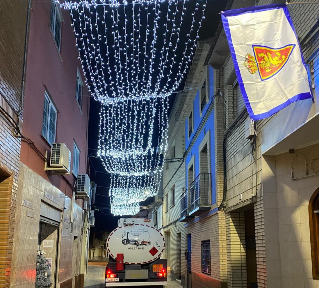 Un árbol de Navidad iluminado junto a un camión blanco con tanque en una calle adoquinada por la noche.