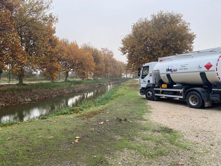 Camión cisterna estacionado cerca de un canal bordeado de árboles con follaje otoñal en un día nublado.