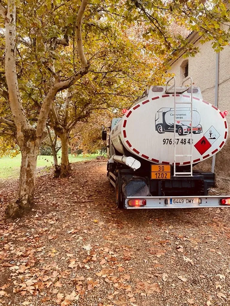 Camión cisterna de combustible estacionado en un camino cubierto de hojas al lado de árboles y un edificio de ladrillos.