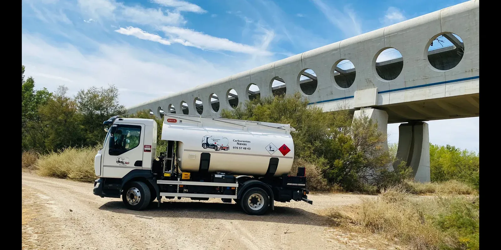 Camión cisterna de combustible blanco en un camino de tierra bajo un puente de hormigón con aberturas circulares. Cielo azul y vegetación al fondo.