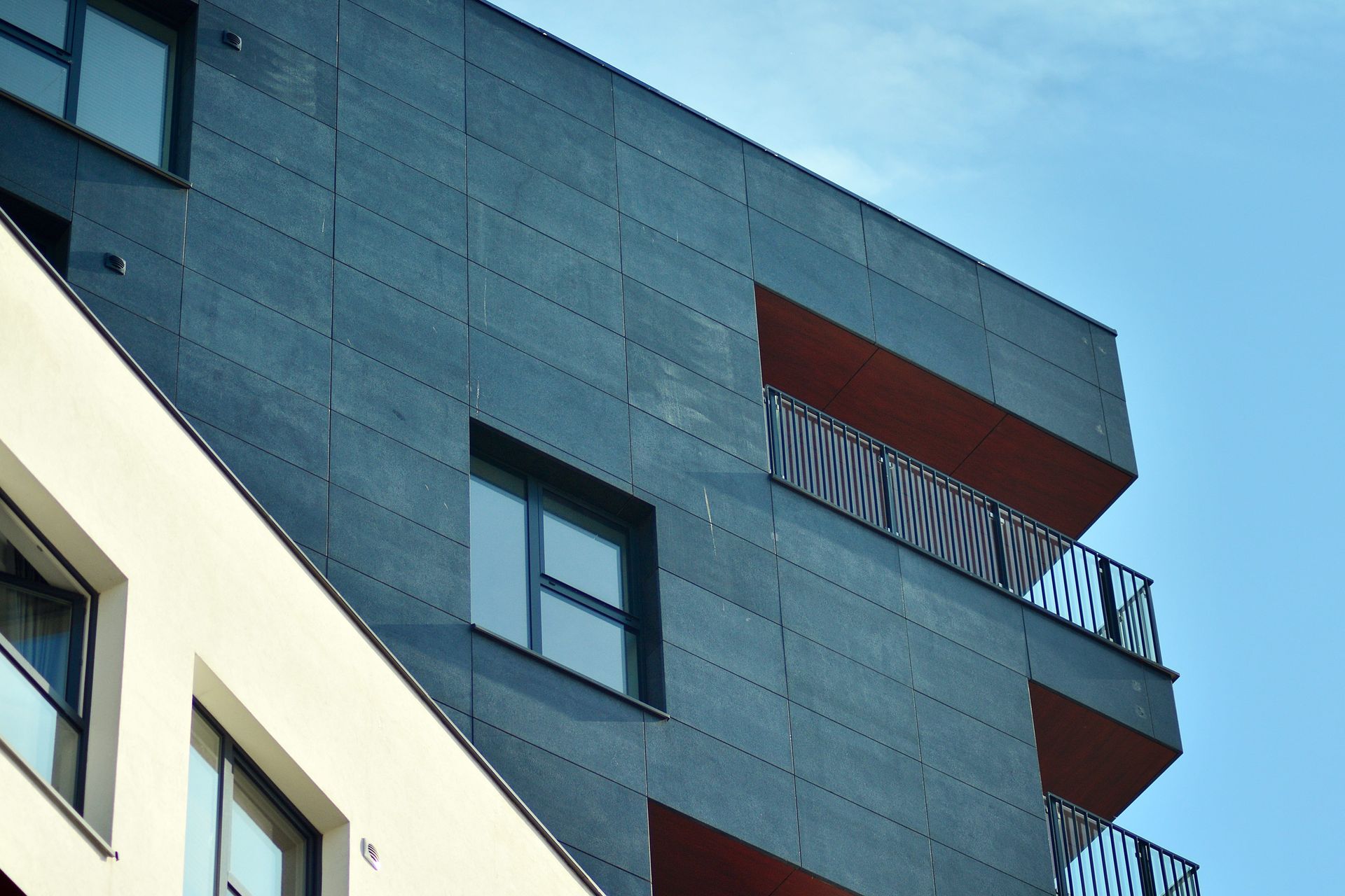 Edificio de apartamentos moderno con fachada gris oscuro, balcones rojos y cielo azul.
