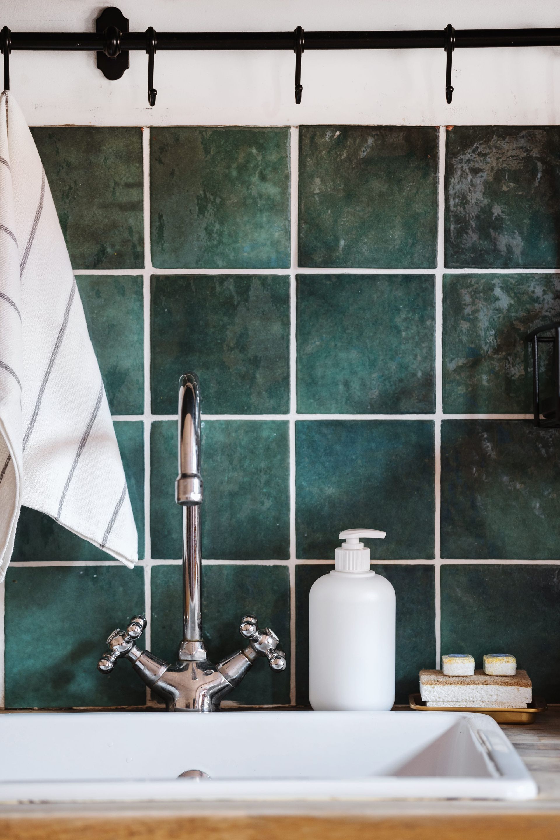Lavabo con grifo cromado, dispensador de jabón blanco, toalla y pared alicatada en verde oscuro.