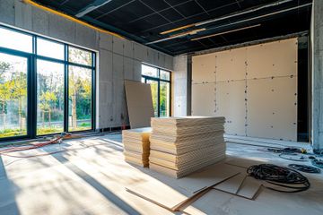 Habitación sin terminar con grandes ventanales, paneles aislantes apilados y cables enrollados en el suelo.