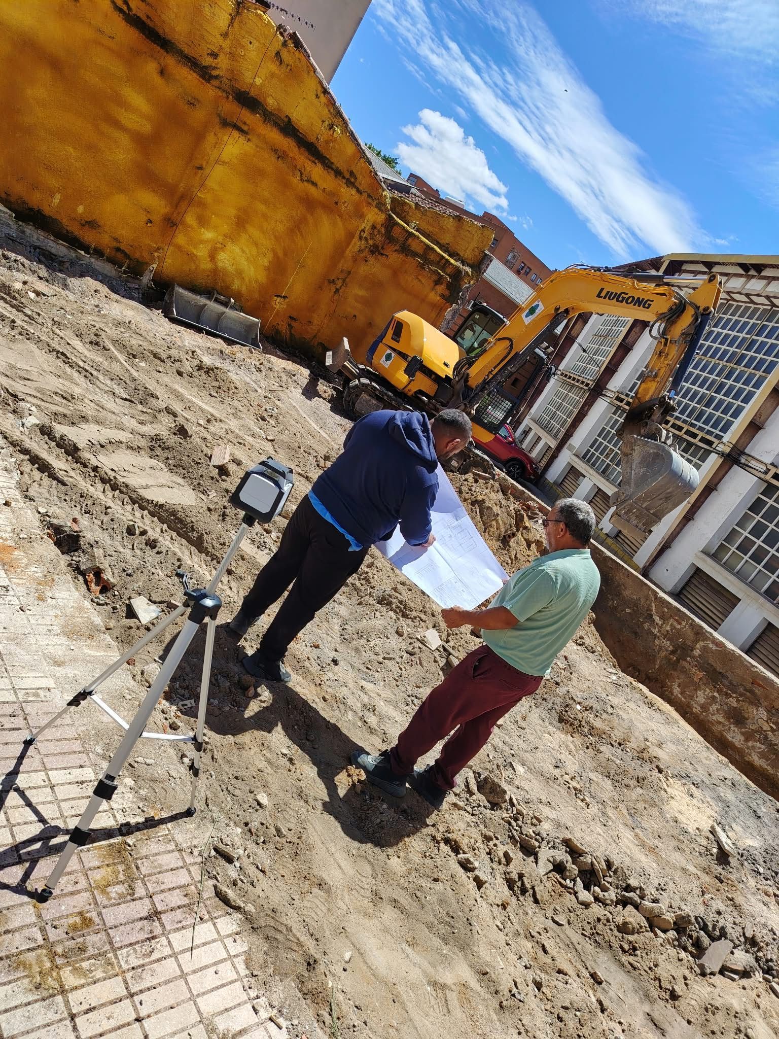 Dos trabajadores en una obra de construcción revisando planos junto a maquinaria pesada y una zona de excavación de tierra.