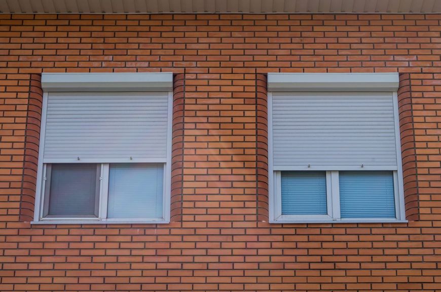 Dos ventanas con persianas blancas en la pared de un edificio de ladrillo rojo.