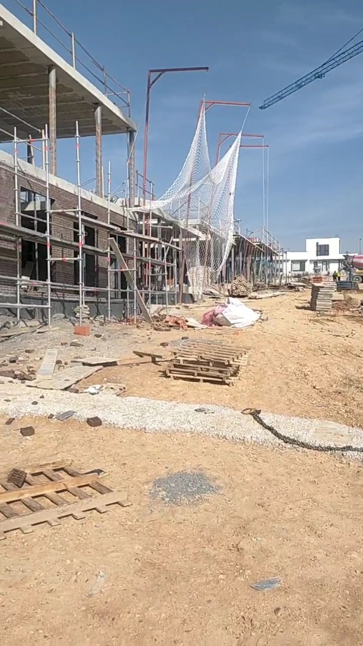 Obra en construcción con andamios, redes y montones de escombros a lo largo de una carretera polvorienta.