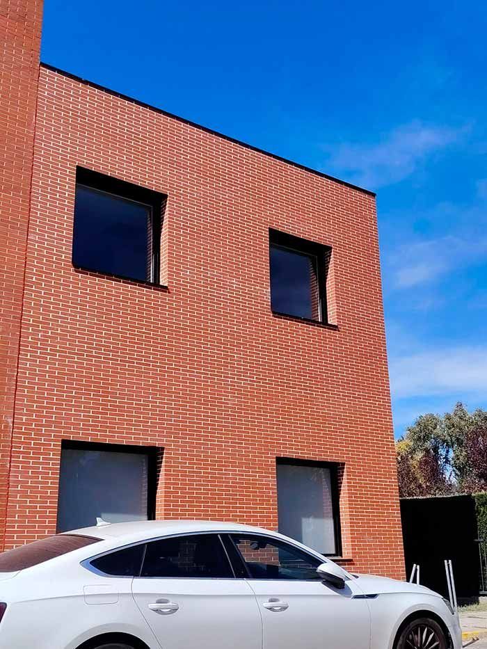 Edificio de ladrillo rojo con ventanas oscuras junto a un coche blanco bajo un cielo azul.