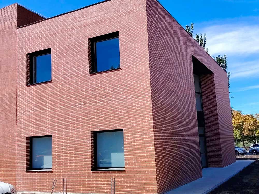 Edificio moderno de ladrillo rojo con ventanas tintadas de azul y una entrada lateral cubierta bajo un cielo despejado.
