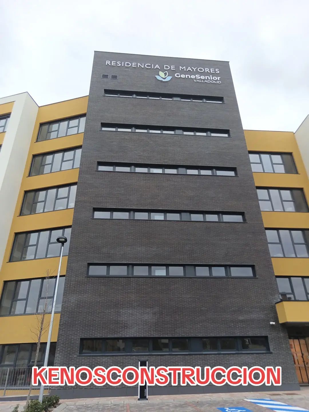 Edificio moderno de varias plantas con fachada oscura y alas laterales amarillas bajo un cielo nublado.
