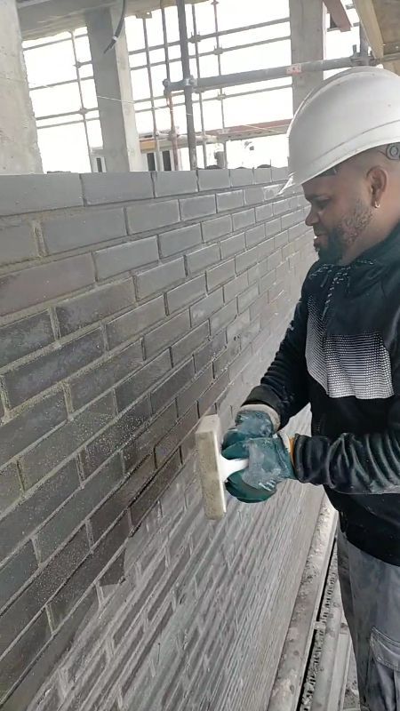 Un trabajador con casco aplica mortero a una pared de ladrillo gris en el interior de una vivienda.