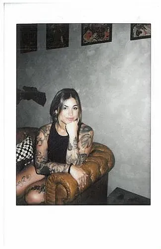 Una mujer con tatuajes está sentada en una silla con la mano en la barbilla.