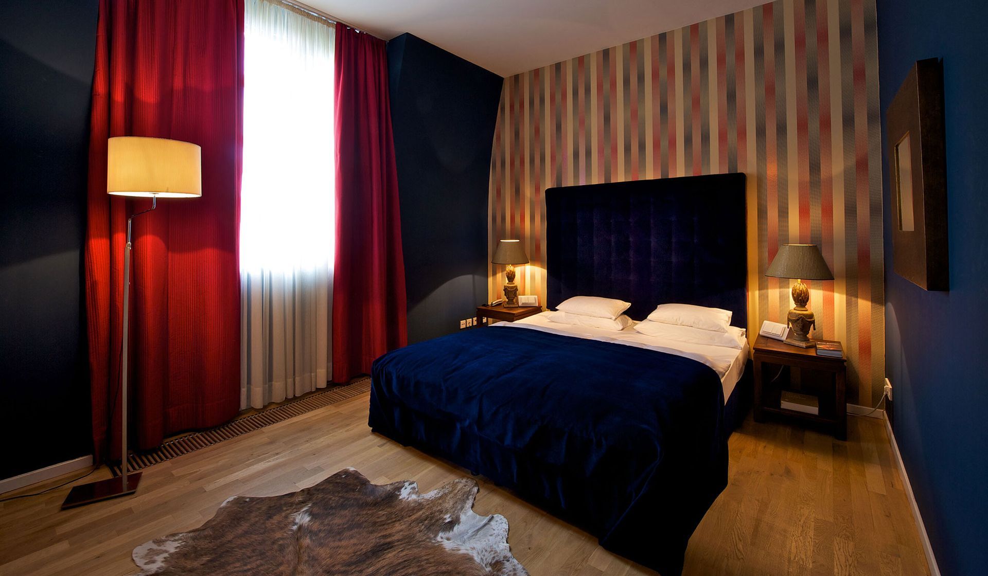 Schlafzimmer mit blauem Samtbett, roten Vorhängen, gemusterter Tapete und Holzboden.