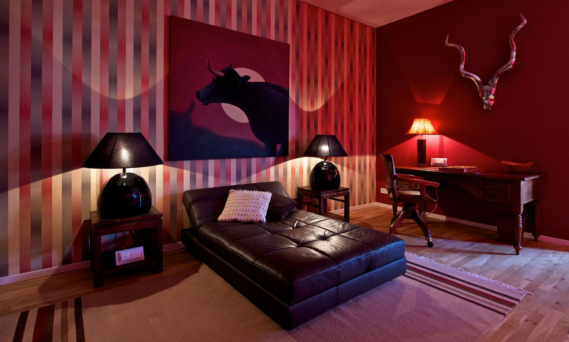 Rotgetöntes Zimmer mit Lederchaiselongue, Kunstwerken und Schreibtisch. Gestreifte Tapete, Lampen und dekorative Tierhörner.