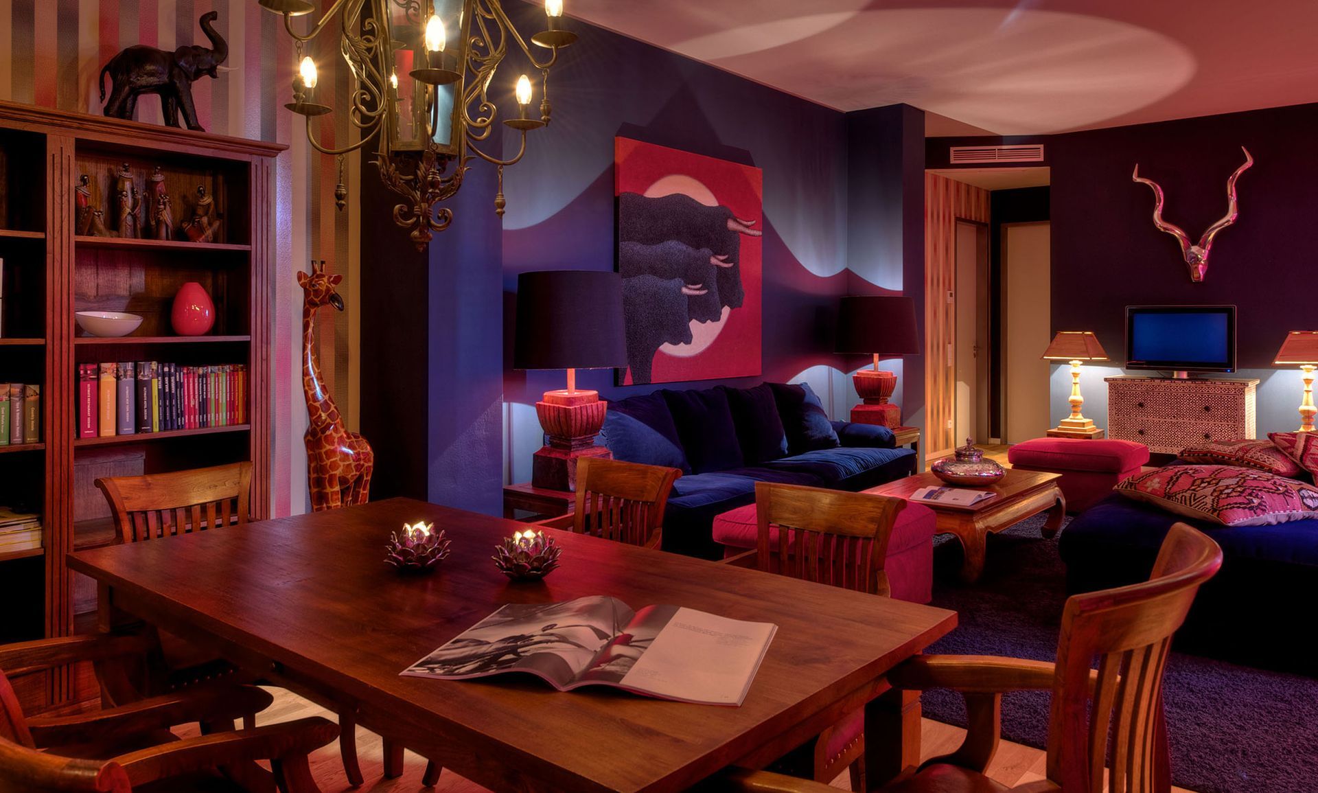 Ein eklektisches Esszimmer mit Bücherregal, Holztisch, Sofa, Kunstwerken und lebhafter violett-roter Beleuchtung.