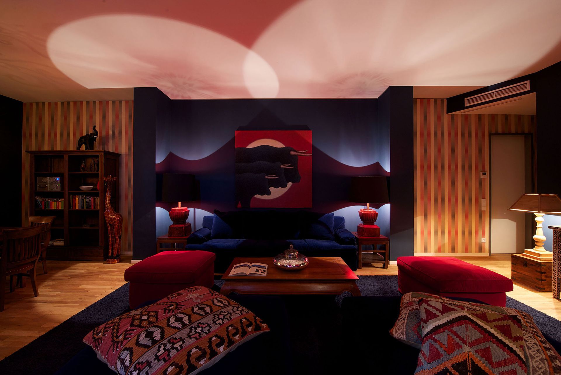 Gemütliches Wohnzimmer mit dunkelblauen Wänden, roten Akzenten und gemusterten Kissen. Sanfte Strahler beleuchten die Decke und die Kunstwerke.
