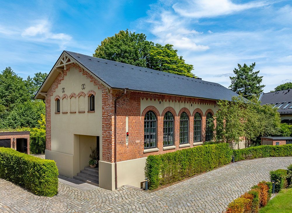 Zweistöckiges Backsteingebäude mit Bogenfenstern, dunklem Dach und einer von Hecken gesäumten Auffahrt.