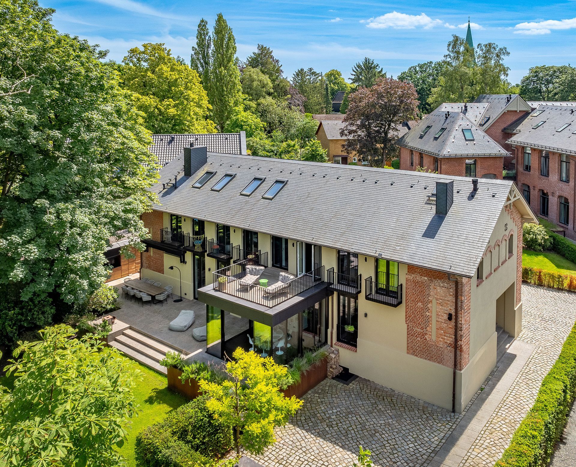 Zweistöckiges modernes Gebäude mit grauem Dach, Ziegel- und hellfarbigen Wänden, umgeben von Bäumen und einer steinernen Auffahrt.