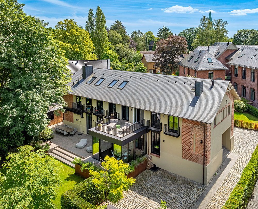 Mehrstöckiges Gebäude mit grauem Dach, Fassade aus Ziegeln und beigem Leder sowie grünem Laubwerk in der Umgebung.
