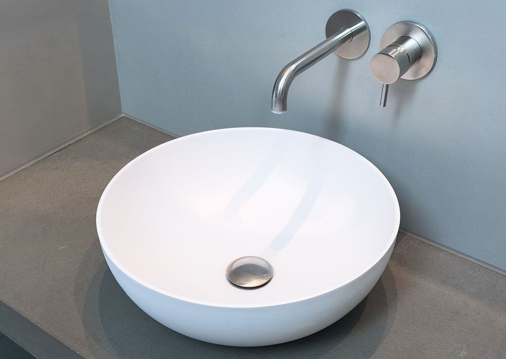 Weißes, rundes Waschbecken auf grauer Arbeitsplatte mit wandmontierter Armatur und Griff.