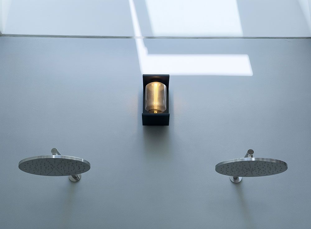 Zwei Duschköpfe flankieren eine Wandleuchte an einer hellgrauen Wand. Natürliches Licht fällt von oben ein.