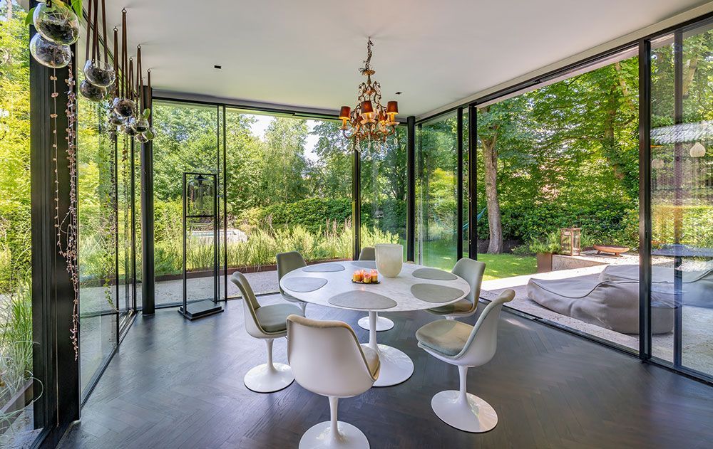 Verglaster Speisesaal mit rundem Tisch, weißen Stühlen und Kronleuchter, mit Blick auf einen üppig grünen Garten.