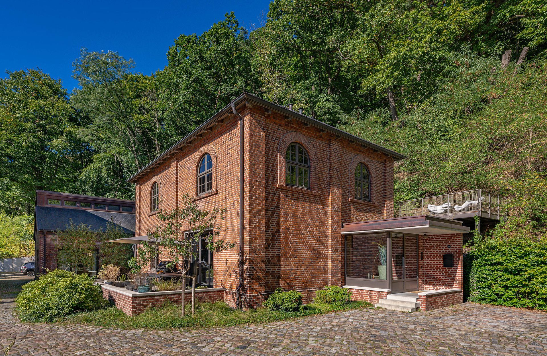 Backsteingebäude mit Bogenfenstern, umgeben von Bäumen. Rechts schließt sich ein kleiner Innenhof an.