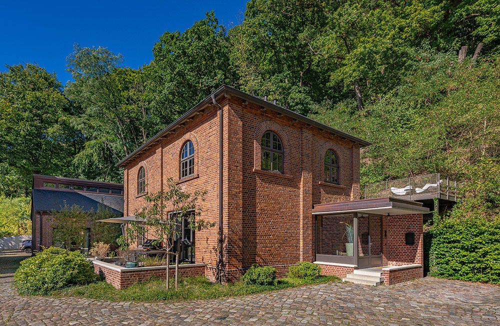 Backsteingebäude mit Bogenfenstern, umgeben von Bäumen. Steinterrasse und moderner Glaseingang.