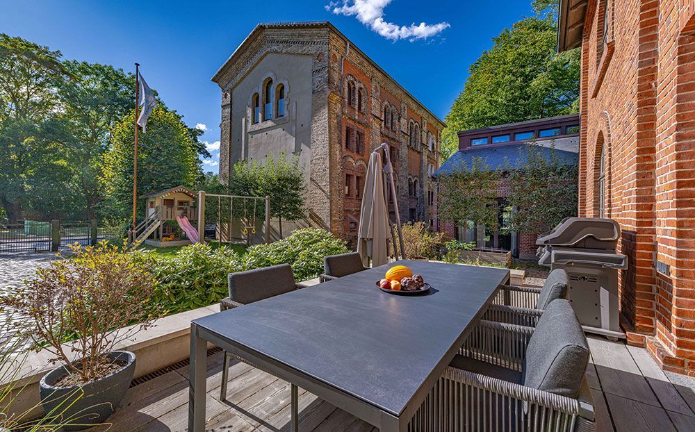 Terrasse mit gedecktem Tisch, im Hintergrund ein rotes Backsteingebäude. Spielplatz in Sicht.