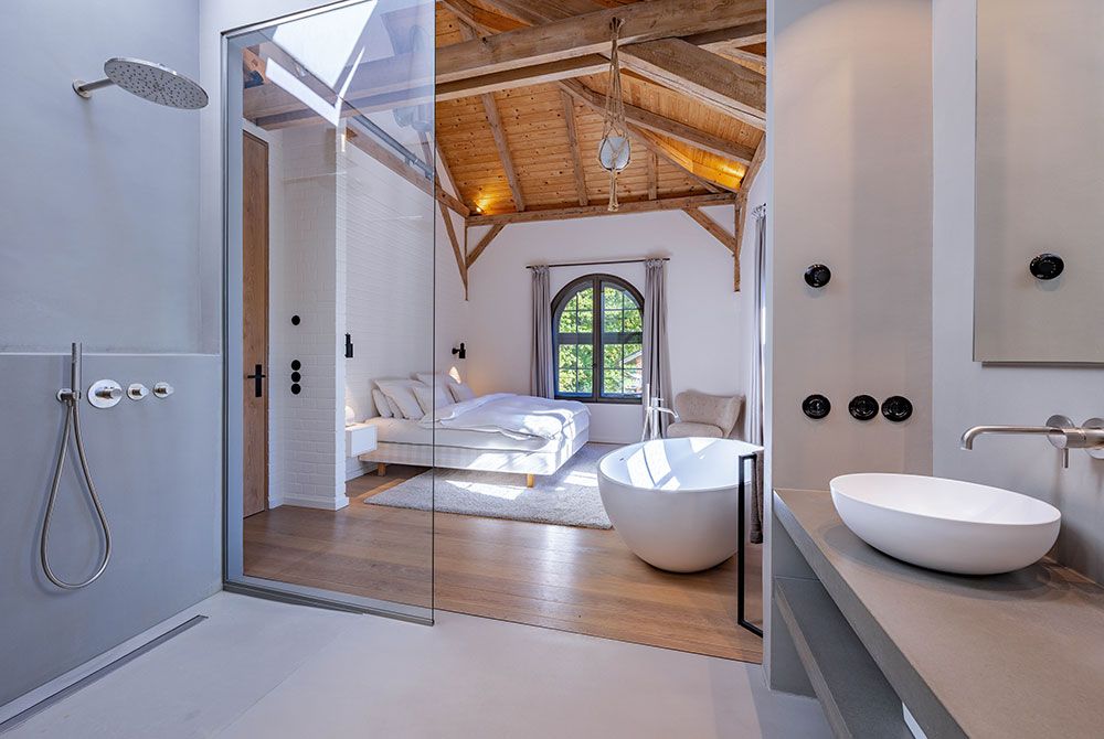 Badezimmer mit Glasdusche, Badewanne, Bett im Hintergrund; helle Wände, Holzbalken.