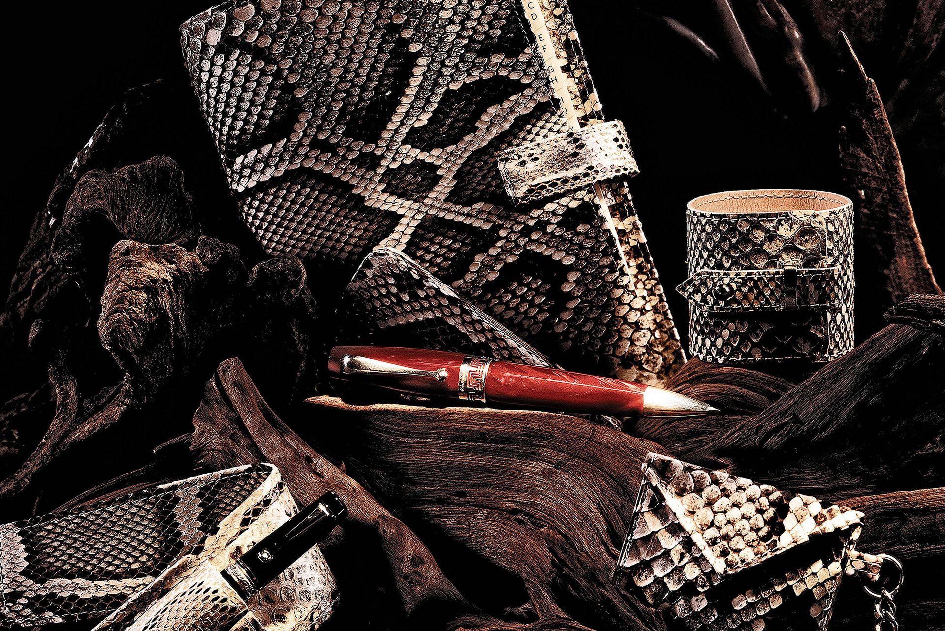 Notizbücher, ein zylinderförmiger Behälter und ein Stift aus Schlangenleder. Alles befindet sich auf braunem Stoff.