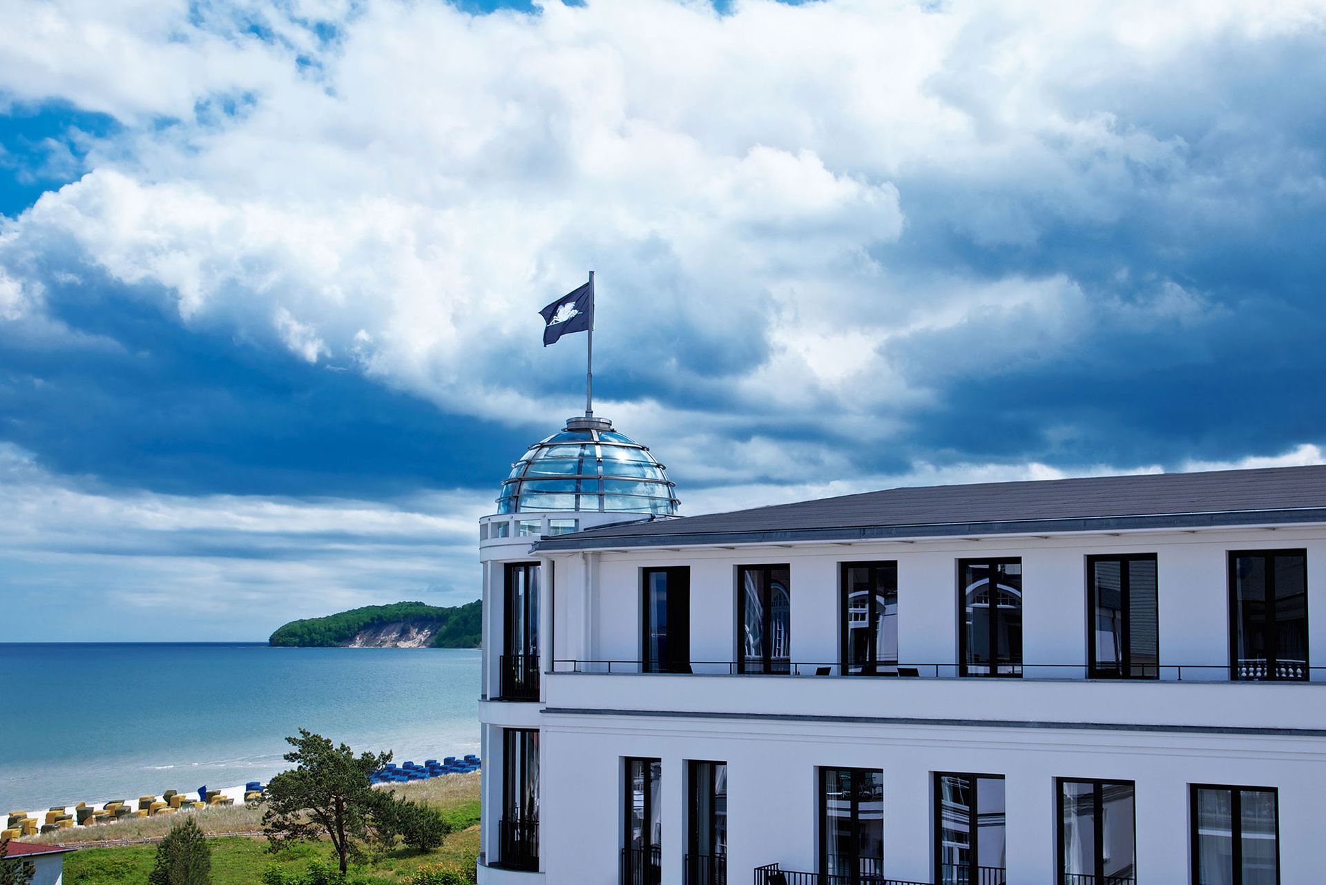 Weißes Gebäude mit Glaskuppel und Flagge, das unter einem bewölkten Himmel auf Strand und Meer blickt.