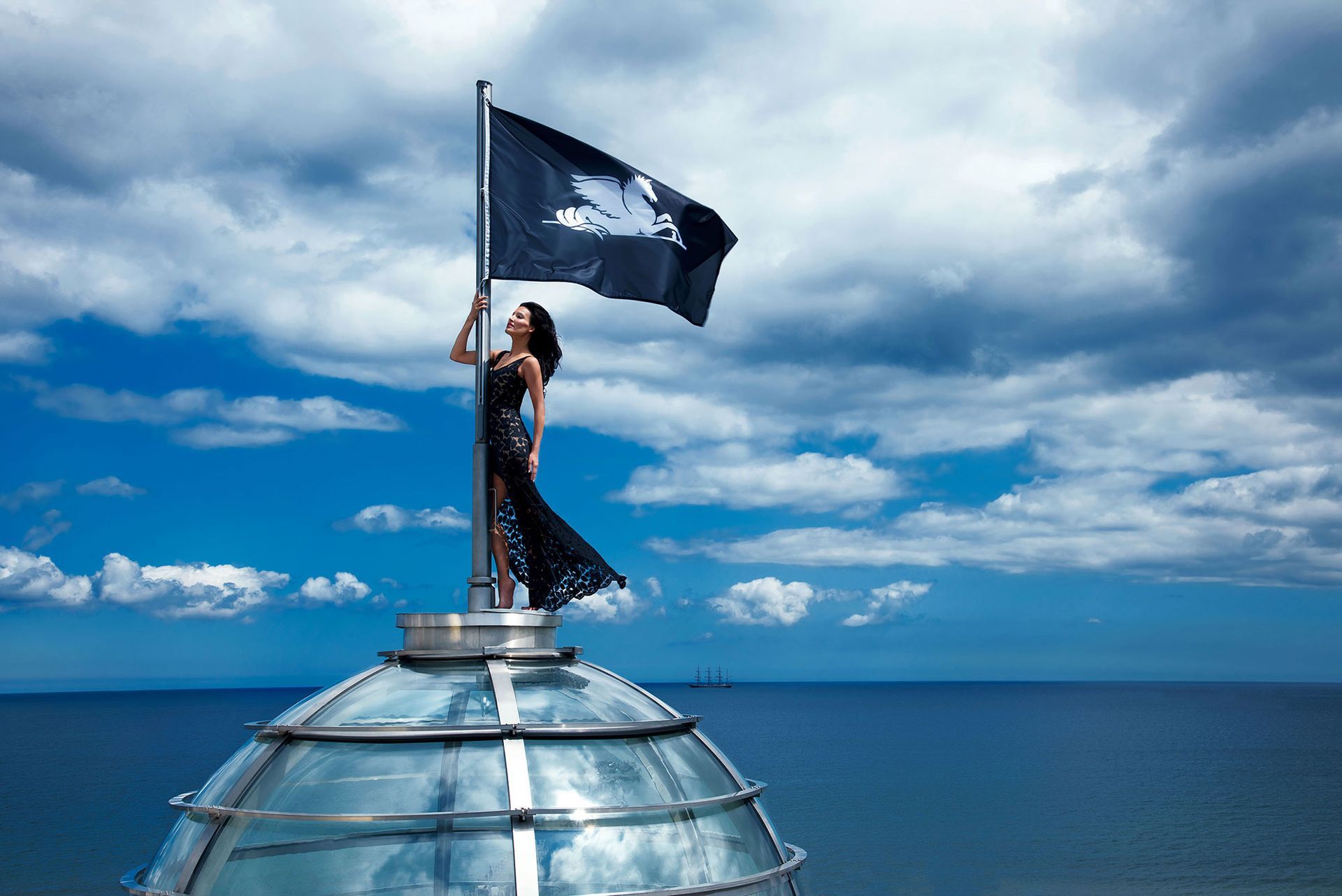 Eine Frau hisst auf einer Glaskuppel eine schwarze Flagge mit weißem Muster; im Hintergrund sind Meer und Himmel zu sehen.