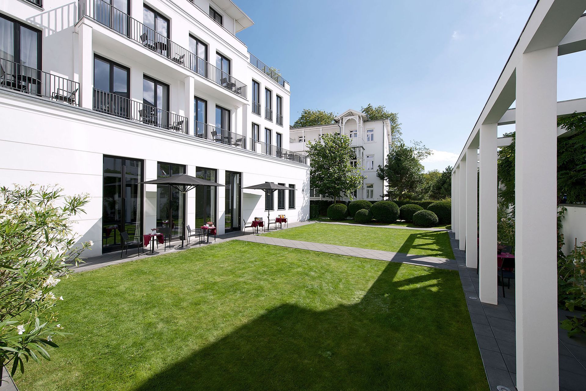 Weißes Hotelgebäude mit Balkonen, die auf einen grünen Rasen und eine Pergola blicken; sonniger Tag.