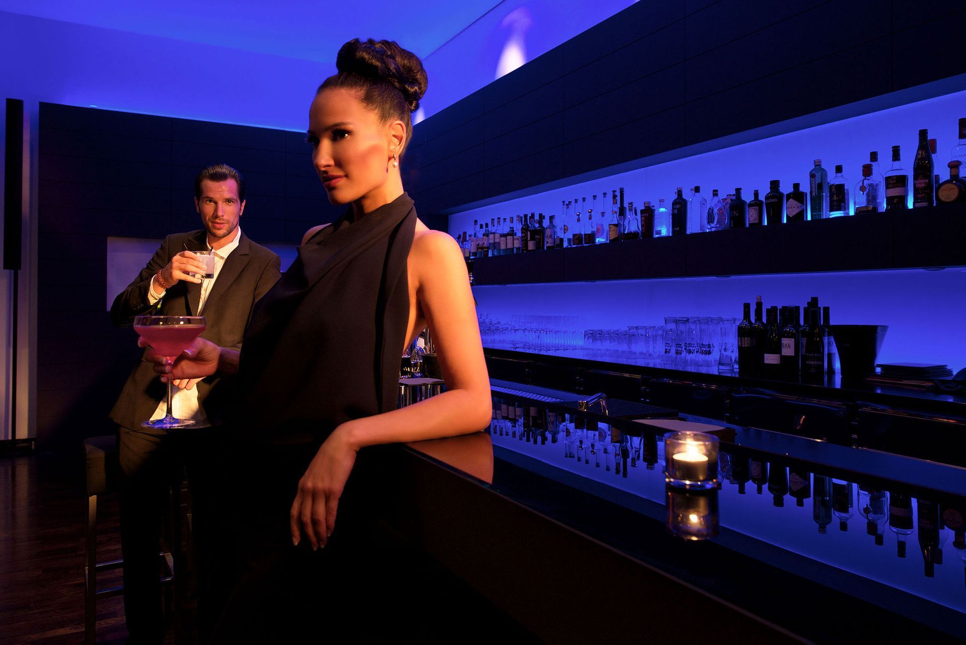 Frau an einer Bar, an den Tresen gelehnt; Mann mixt hinter ihr einen rosa Drink; blaue Beleuchtung; Flaschen in Regalen.