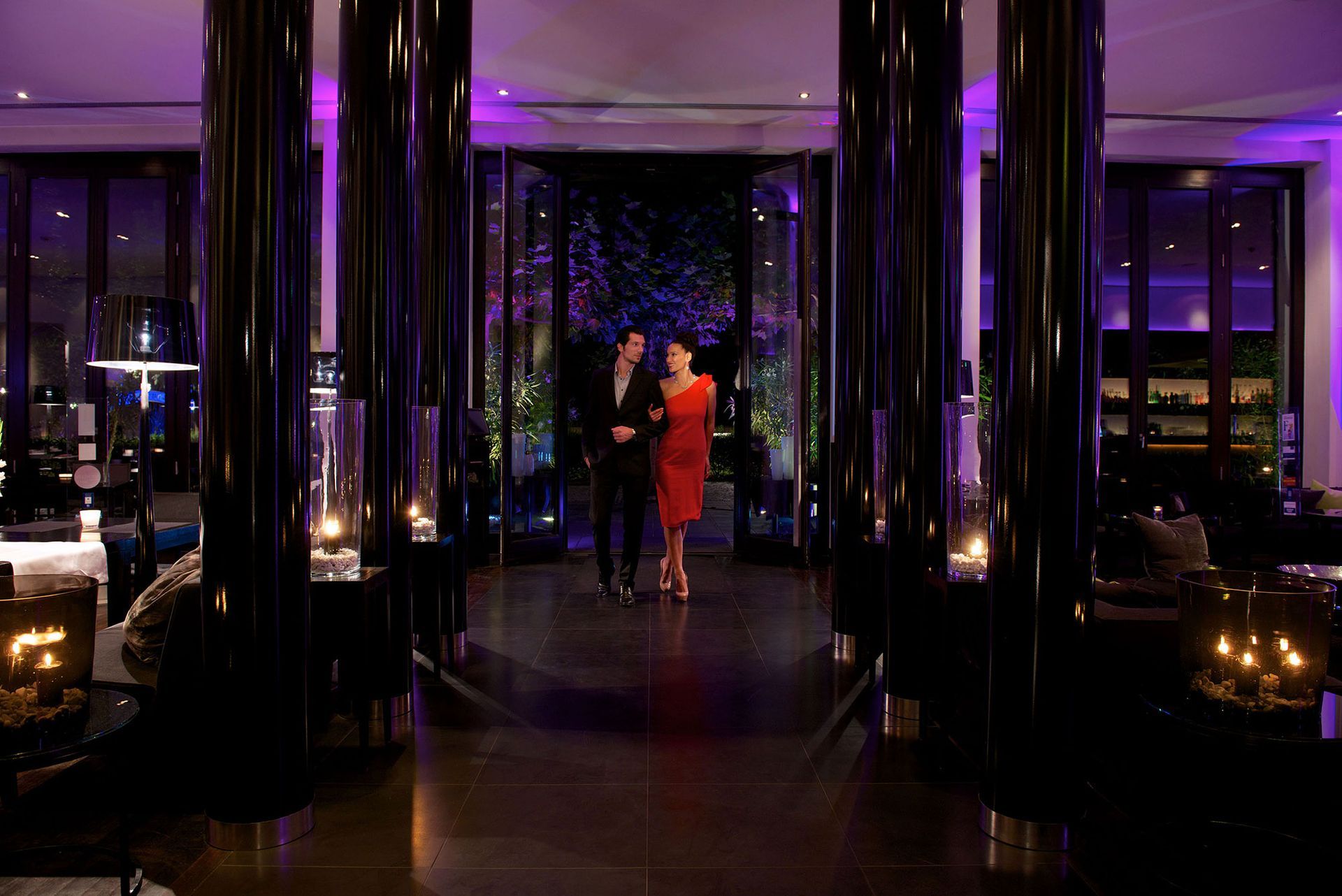 Ein Paar betritt Arm in Arm eine schwach beleuchtete, moderne Lounge mit dunklen Säulen und violetter Beleuchtung.