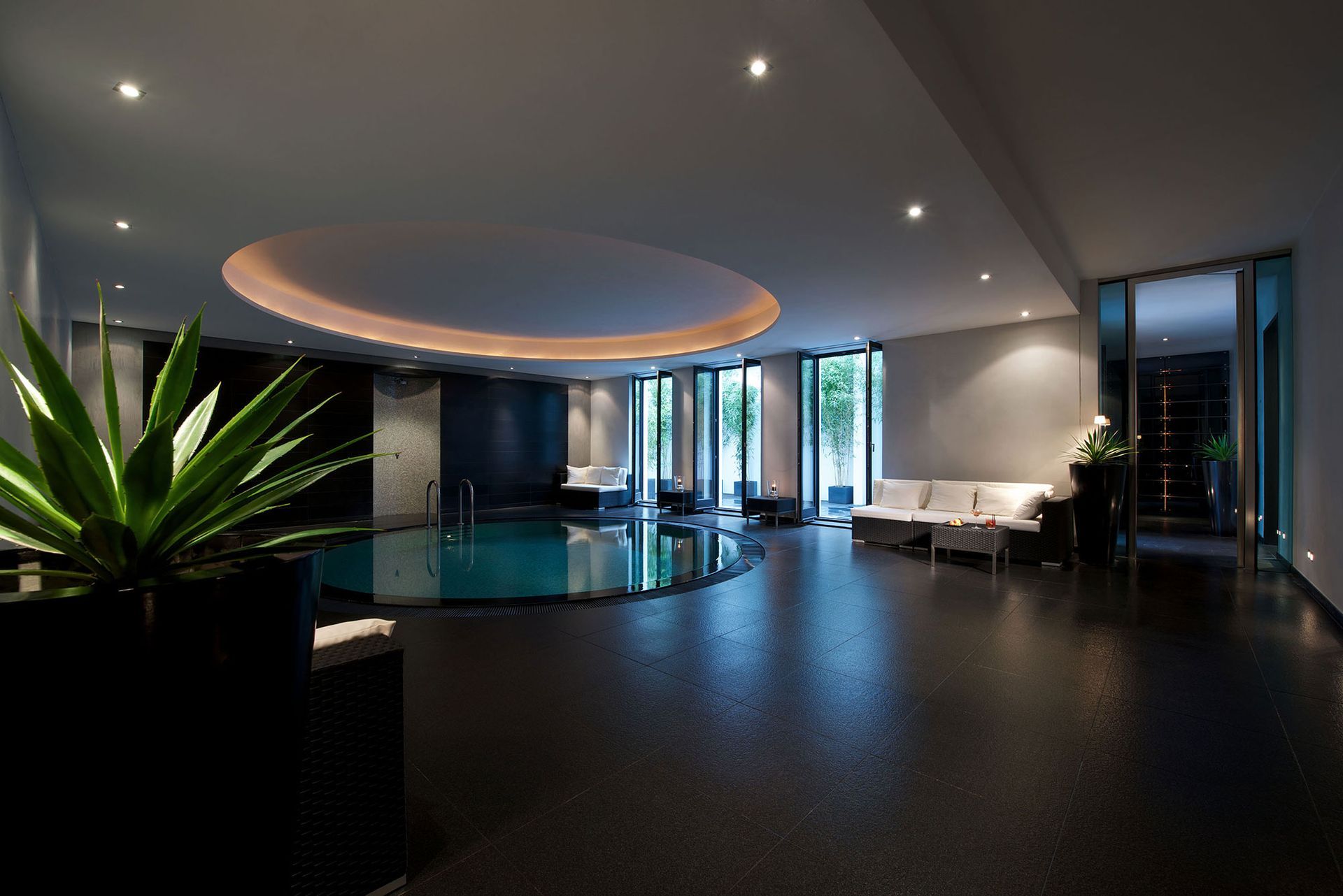 Hallenbad mit schwarzem Boden, runder Deckenleuchte und bodentiefen Glastüren.