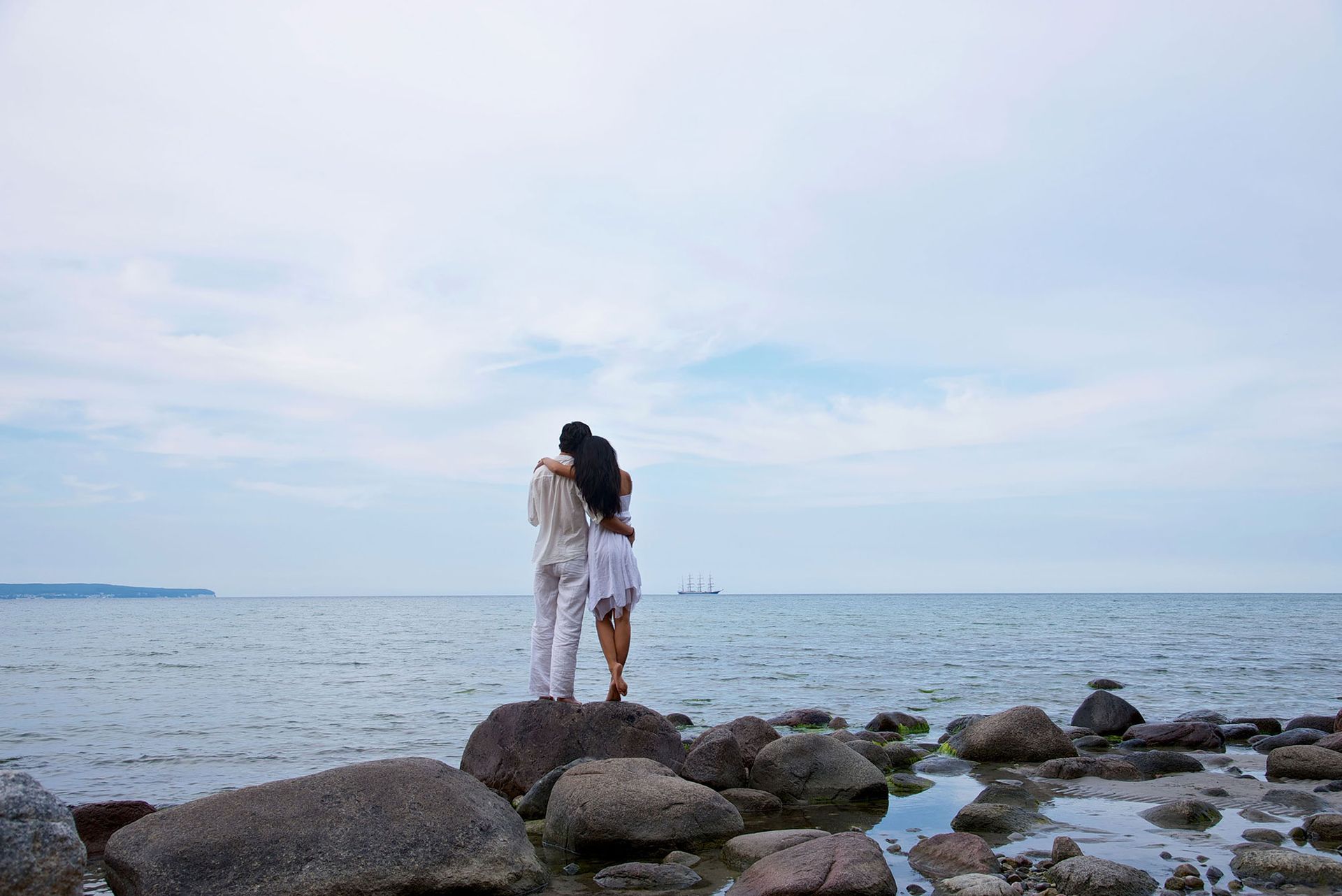 Ein Paar umarmt sich an einem felsigen Ufer und blickt unter einem bewölkten Himmel aufs Meer hinaus.