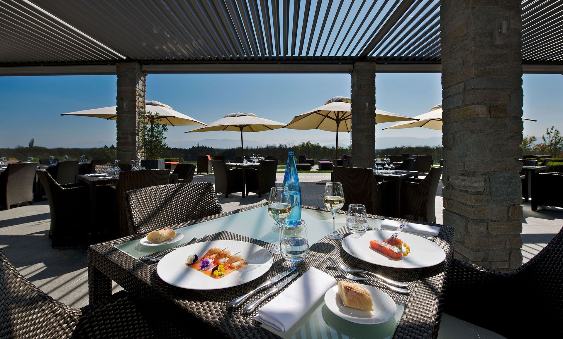 Tischdekoration in einem Restaurant im Freien mit Speisen und Getränken; Blick auf Sonnenschirme und die Landschaft in der Ferne.