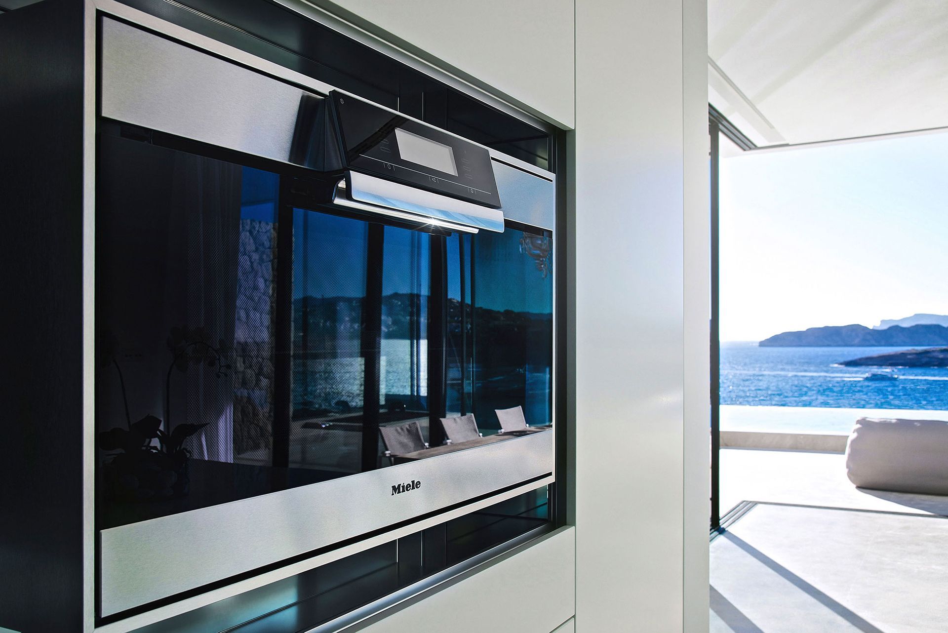Eingebaute Mikrowelle mit verspiegelter Glastür. Durch die offene Tür eröffnet sich ein Blick auf das Meer, die Berge und die Terrassenmöbel.