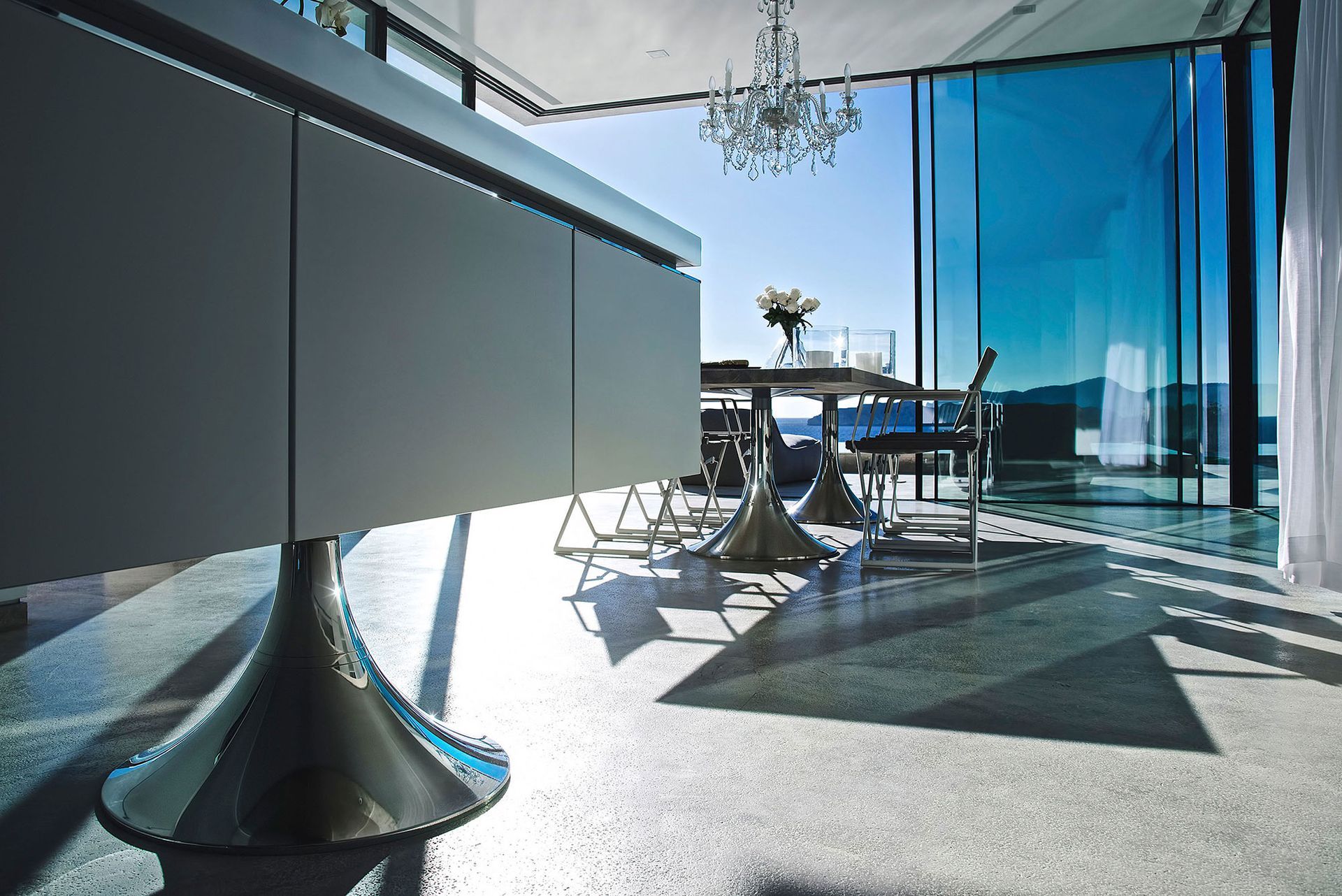 Modernes Interieur mit Glaswänden, Tisch, Kronleuchter und Meerblick.