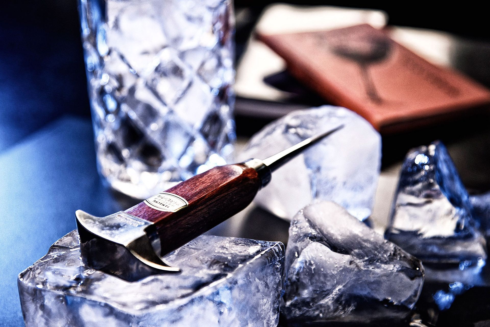 Ein Eispickel ruht auf Eiswürfeln, im Hintergrund ist ein Glas mit Eis zu sehen.