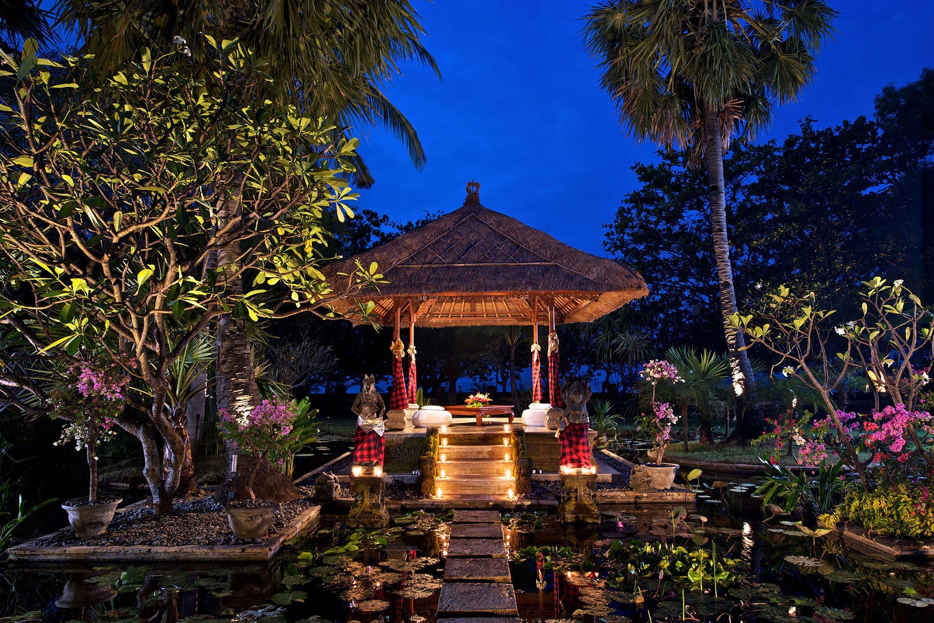 Nachts ein beleuchteter Pavillon über dem Wasser; üppiges Laubwerk, Kerzen und tropische Bäume.